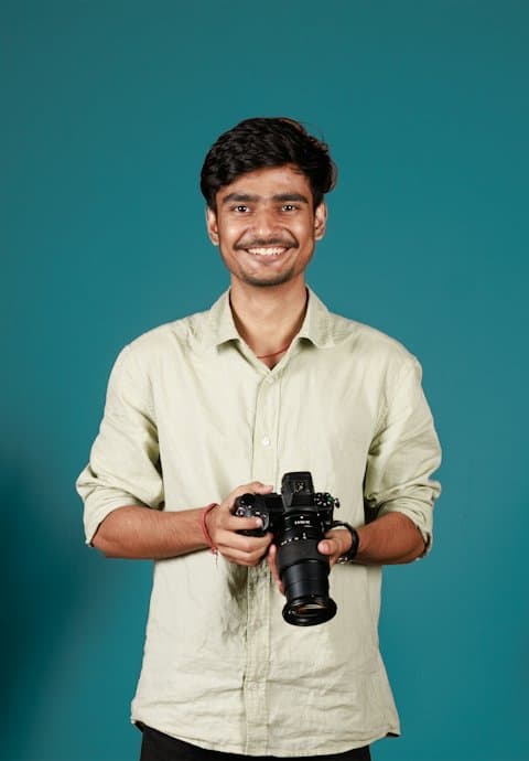 A smiling young man in a light green shirt holding a mirrorless camera in front of a teal studio wall