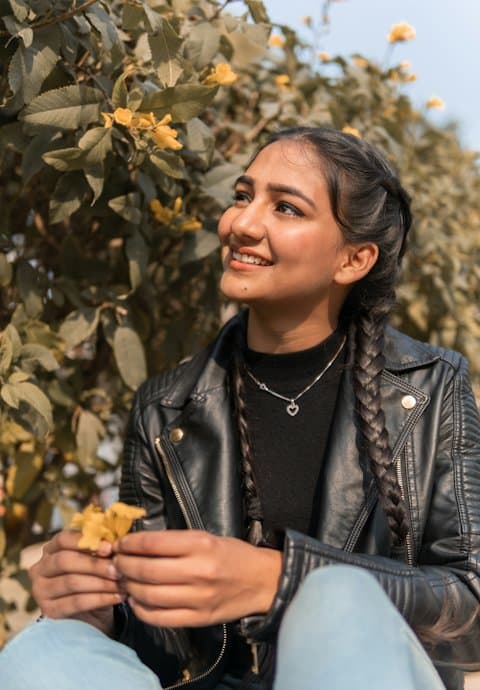 A smiling young woman with long braids and a black leather jacket holding yellow flowers in front of a flowering bush