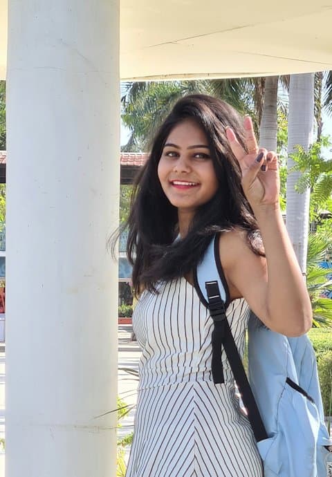 A smiling college student with a light blue backpack waving a peace sign beside a white pillar on a sunny campus