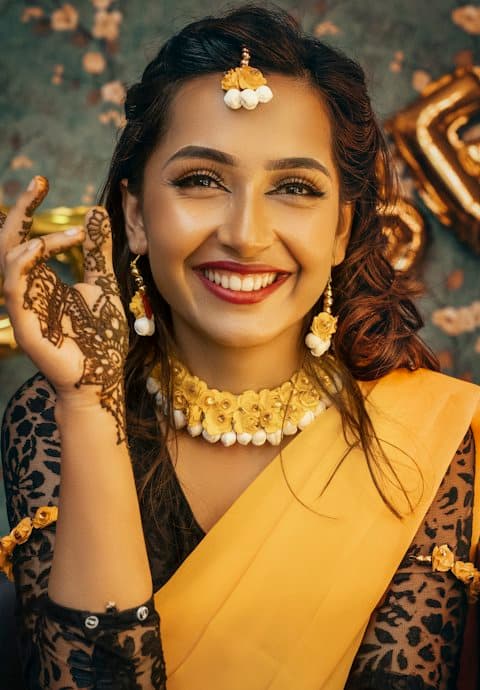 A smiling woman in a mustard saree with a floral flower necklace, maang tikka and mehendi on her raised hand, gold balloons behind her