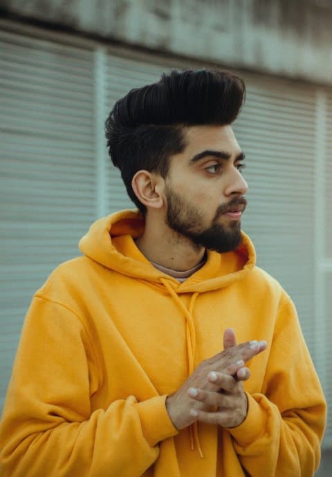 A young bearded man in a mustard yellow hoodie looking to the side, hands clasped, in front of a grey shutter