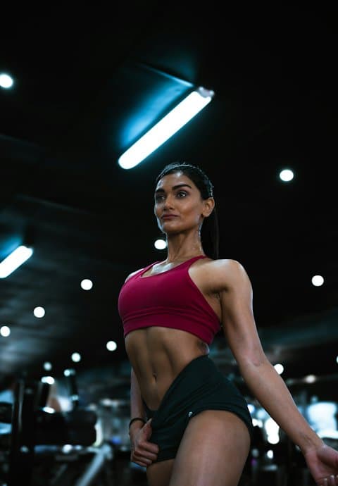 A fitness coach in a red sports top and black shorts standing in a dark gym under bright ceiling lights