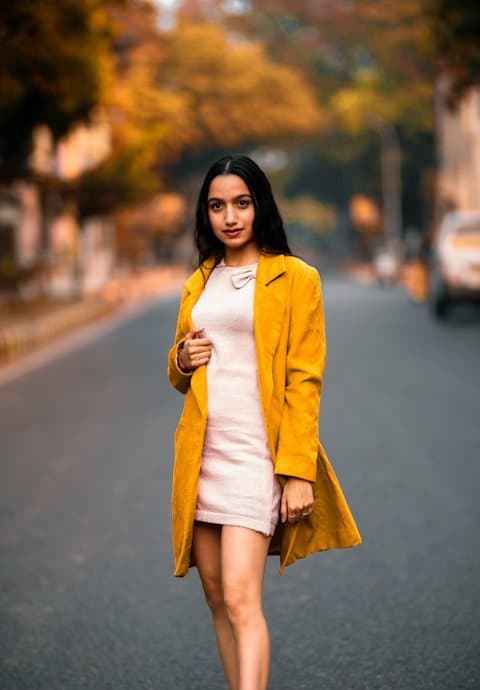 A young woman in a mustard yellow coat over a pale pink dress walking down an empty tree-lined road in autumn