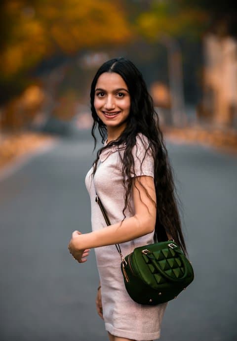 A smiling young woman with long dark hair in a pale pink dress, carrying a green quilted handbag on a tree-lined road