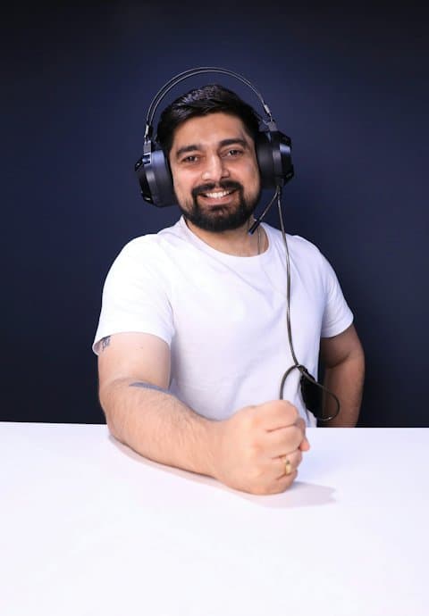 A smiling bearded man in a white T-shirt wearing a black headset with a microphone, sitting at a white desk against a dark blue wall