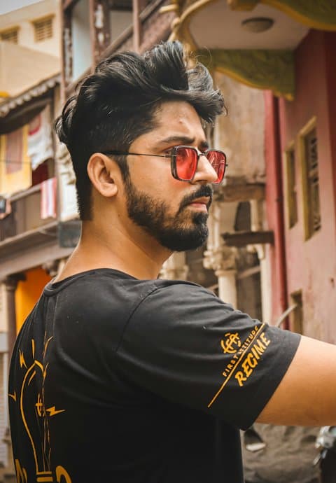 A bearded creator in red-tinted sunglasses and a black tee looking over his shoulder on an old city street