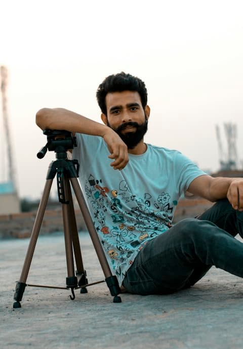 A bearded man in a doodle print tee sitting on a rooftop at dusk, resting his arm on a camera tripod