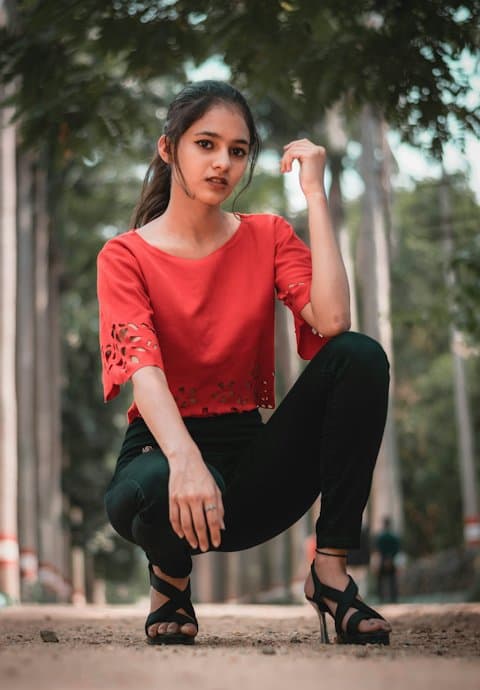 A young woman in a red cutwork top and black jeans crouching on a tree-lined dirt path, looking at the camera