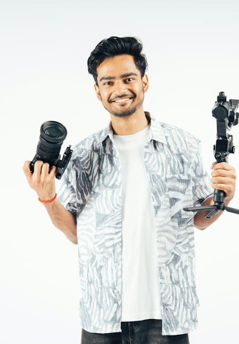 A smiling young man in a printed shirt holding a camera in one hand and a gimbal in the other against a white wall