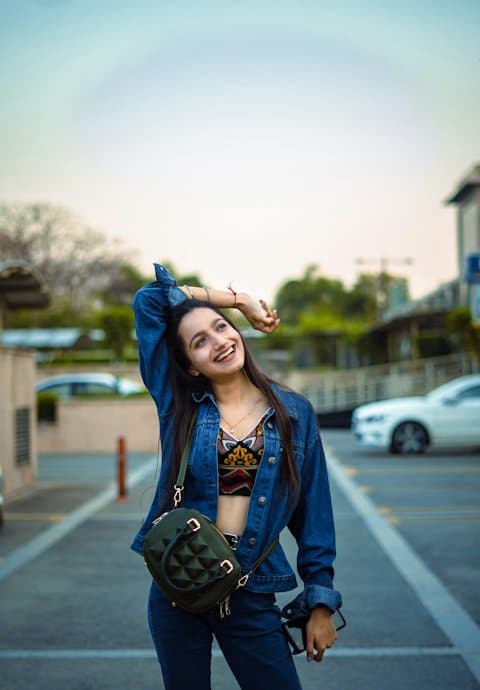 A laughing young woman in a denim jacket with a small green shoulder bag, one arm raised, in an open parking lot at sunset
