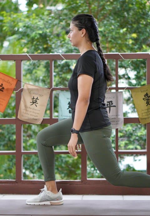 A yoga creator in a black tee and olive leggings holding a low lunge on a balcony hung with prayer flags